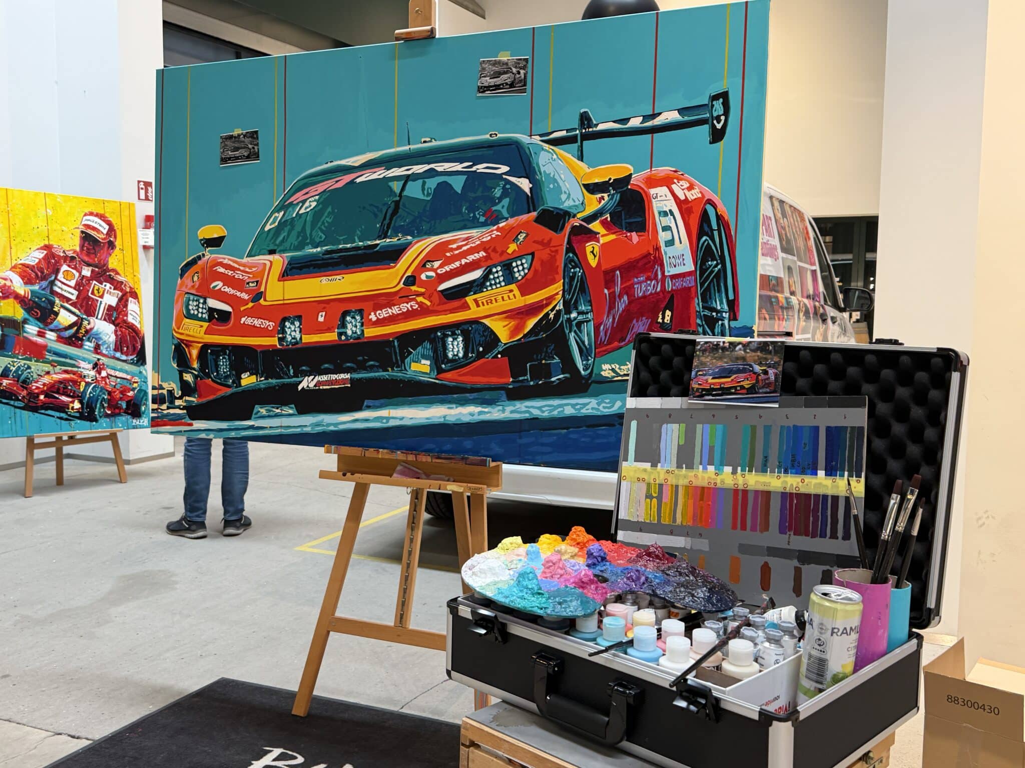 Large vibrant painting of a red Ferrari race car on an easel in an artist studio, with paints and color swatches nearby.