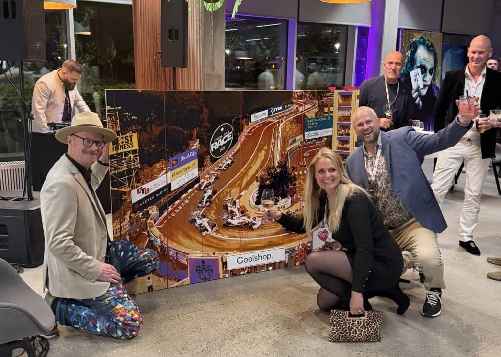 Group of people posing indoors beside a large 'Cool Car Race 2026' race-track backdrop, some holding drinks.