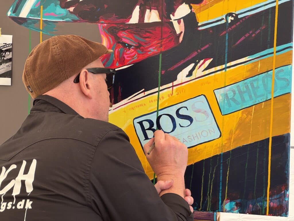 Person wearing a brown corduroy cap and sunglasses painting a bright, abstract mural on a wall with bold yellow, blue, and orange panels, seen from behind.