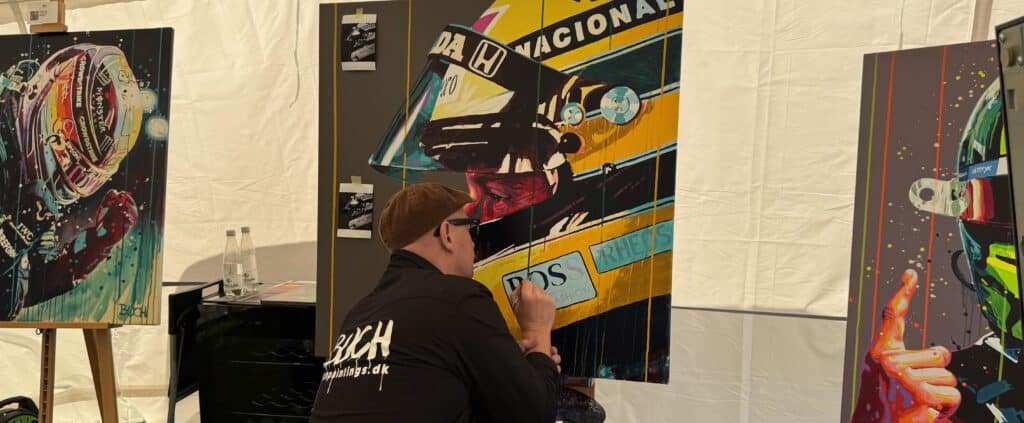 Artist in a brown cap painting a tall colorful mural panel in a tented space, seen from behind with water bottles and art in the background