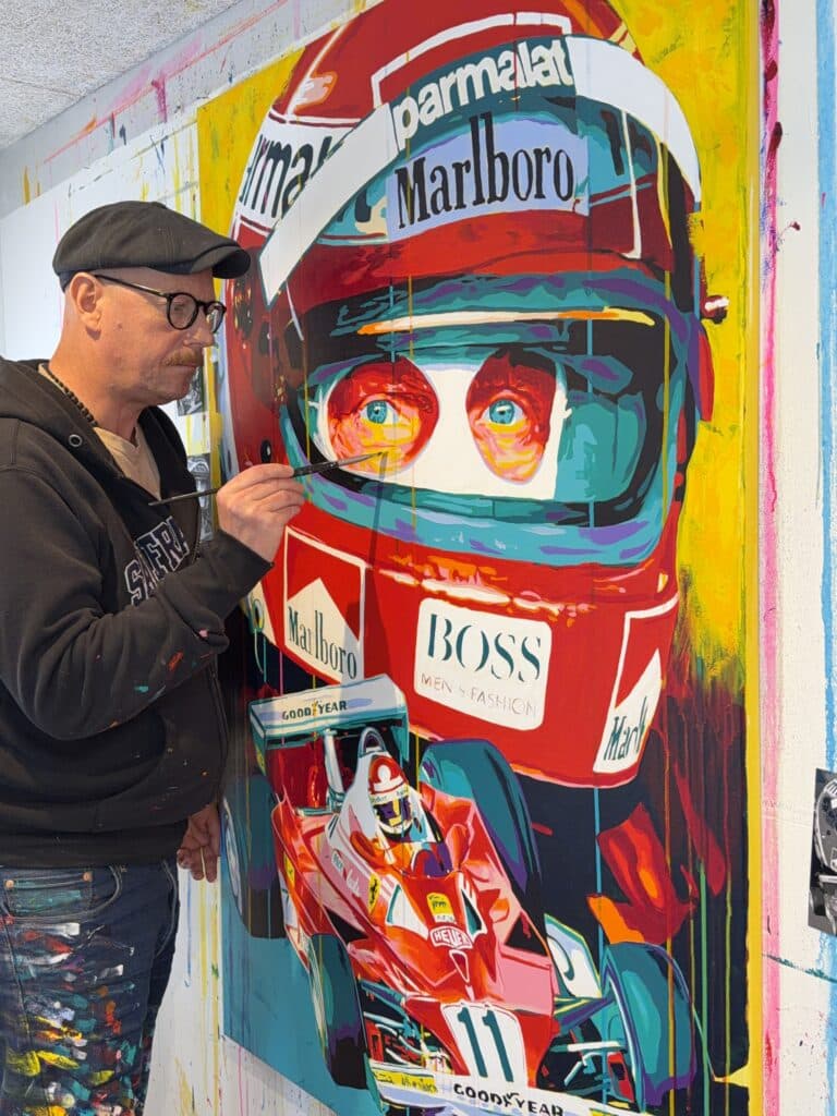 Artist wearing a cap and glasses paints a bright, oversized racing-helmet mural on a wall, with Marlboro and Goodyear logos visible.
