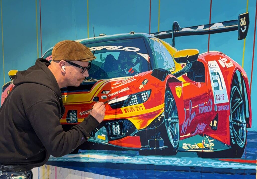 Man in a brown cap and hoodie paints a bright red Ferrari race car mural on a blue wall with paint-splattered pants nearby.