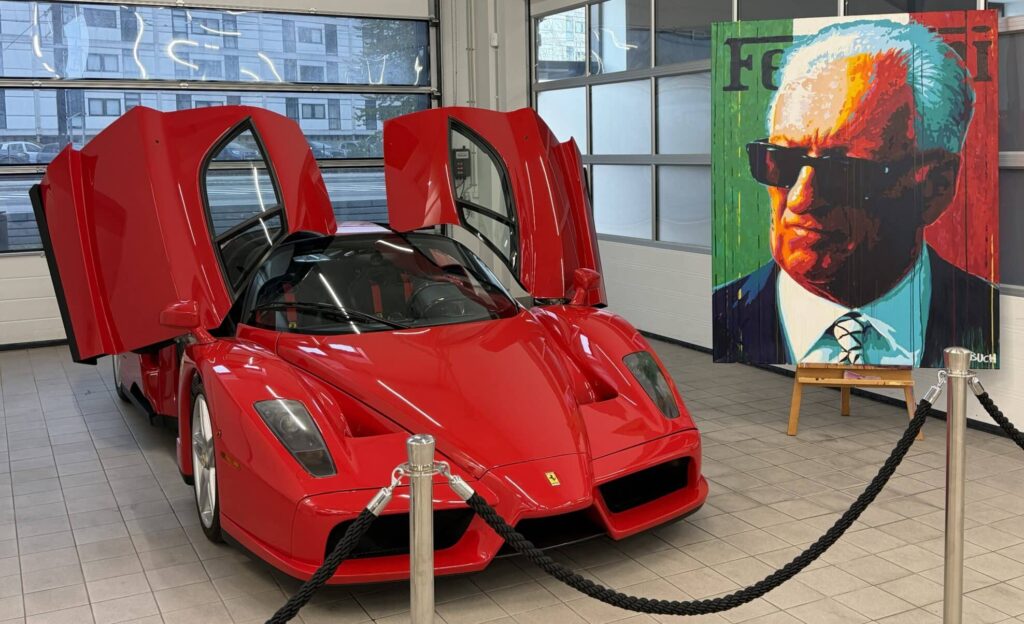 Red Ferrari with gull-wing doors open on a showroom floor beside a colorful portrait panel.